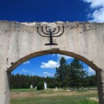Zwietrzały betonowy łuk z symbolem czarnej menorah na szczycie stoi na trawiastym polu, wśród drzew i błękitnego nieba z chmurami w tle.