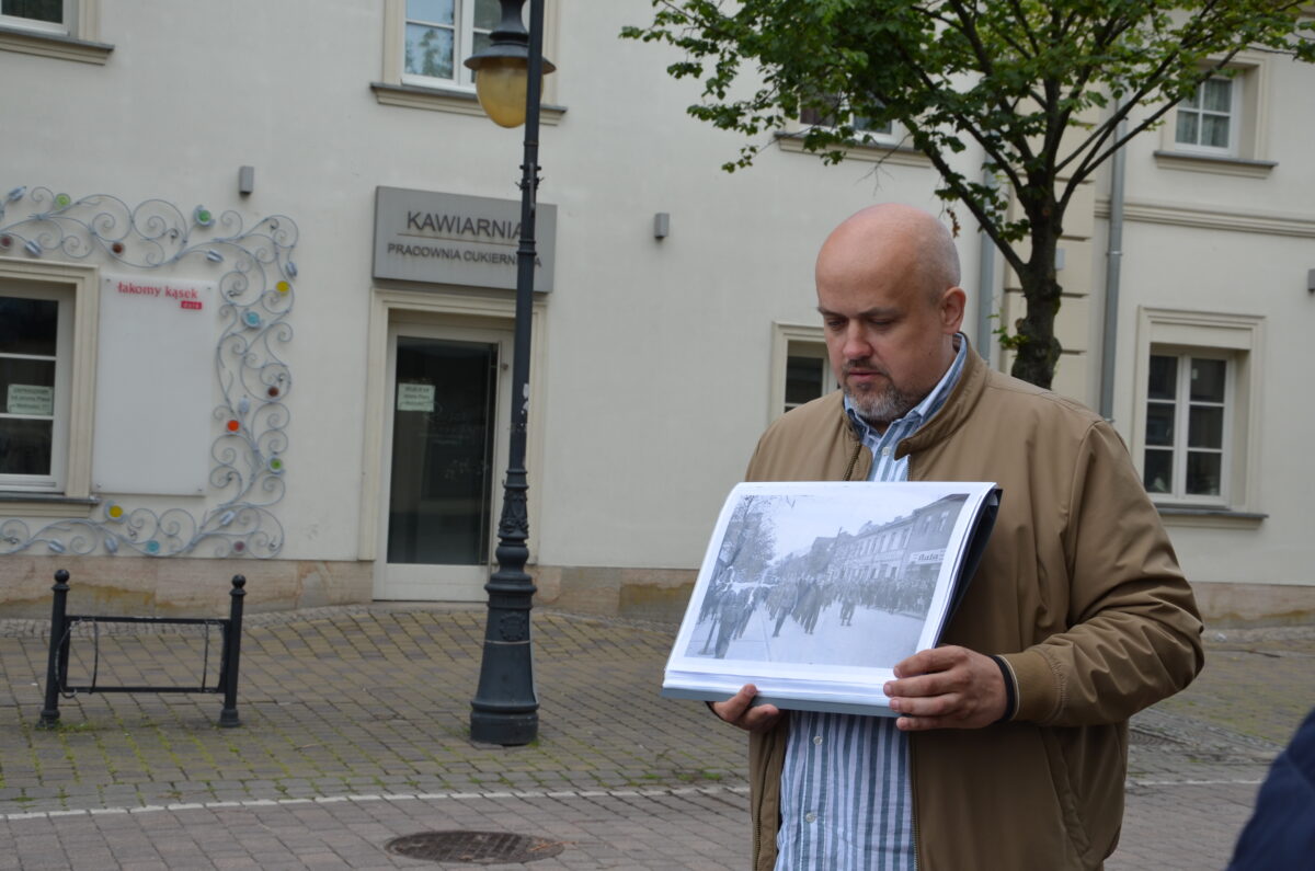 Łukasz Daniewski stoi na brukowanej ulicy, trzymając duże czarno-białe zdjęcie, na tle budynków, drzewa i latarni. Na budynku widnieje napis Kawiarnia Pracownia Cukiernicza.