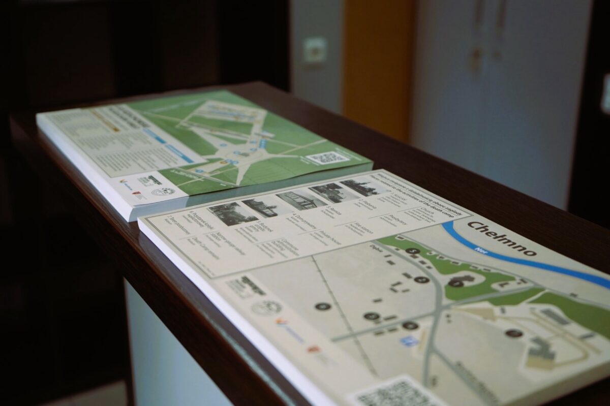 Dwie broszury informacyjne z mapami i tekstem są umieszczone na ciemnym drewnianym blacie. Jedna broszura nosi tytuł „Chelmno” i pokazuje mapę z opisanymi lokalizacjami i małymi obrazkami. Tło jest lekko nieostre.