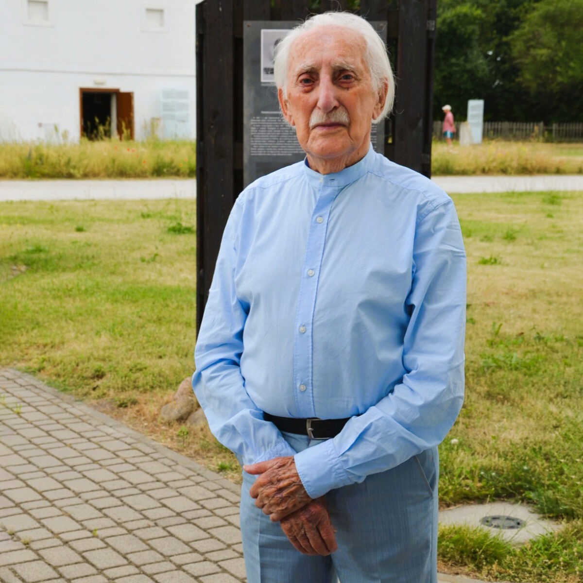 Starszy mężczyzna z białymi włosami i wąsami stoi na zewnątrz na brukowanym chodniku., ubrany w jasnoniebieską koszulę zapinaną na guziki. Znajduje się przed ciemną tablicą informacyjną i trawiastym obszarem, z budynkiem i drzewami w tle.