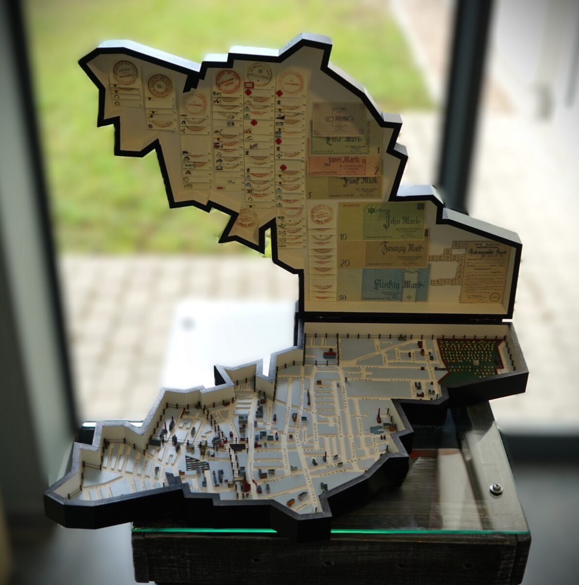 Model 3D w kształcie łódzkiego getta, z górną połową otwartą, aby odsłonić mapy, diagramy i małe figurki w środku, wystawiony na stojaku w pobliżu dużego okna z rozmytym widokiem na zewnątrz.