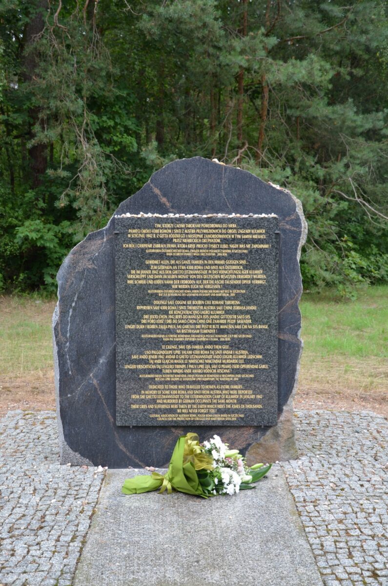 Tablica upamiętniająca z ciemnego kamienia z inskrypcjami stoi na zewnątrz na kamiennej platformie, otoczona drzewami. U podstawy pomnika leży bukiet białych kwiatów zawiniętych w zielony papier.
