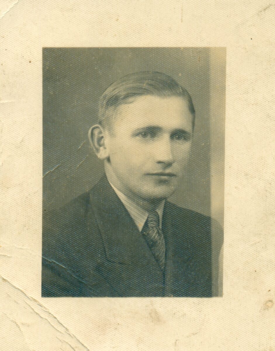 Utrzymany w tonacji sepii, zabytkowy portret młodego mężczyzny w garniturze i krawacie, z krótkimi, starannie zaczesanymi włosami, pozującego na prostym tle. Zdjęcie jest lekko zużyte i postarzone.