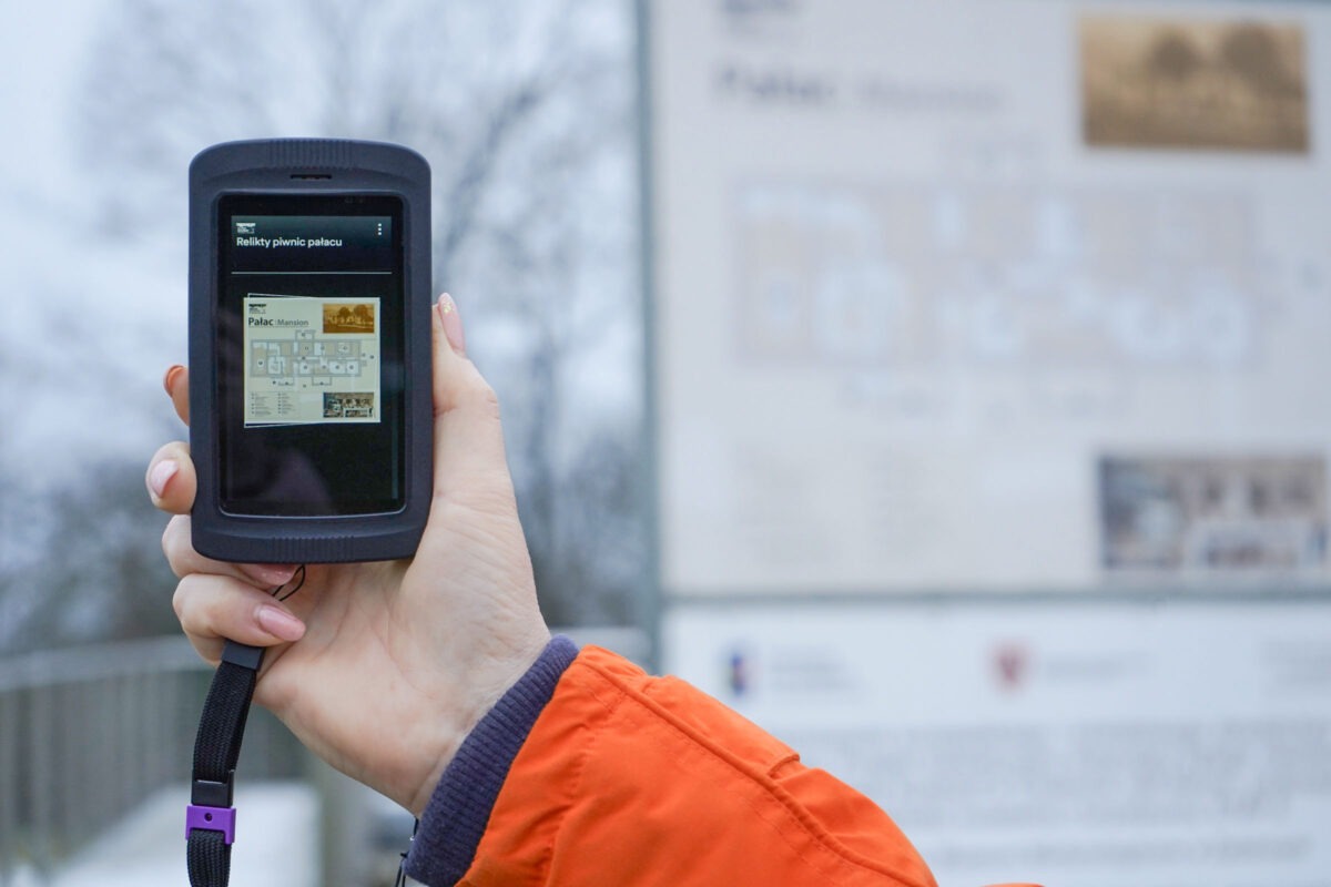 Osoba w pomarańczowej kurtce trzyma ręczne urządzenie wyświetlające cyfrową wersję tablicy informacyjnej, która jest widoczna niewyraźnie w tle na zewnątrz.