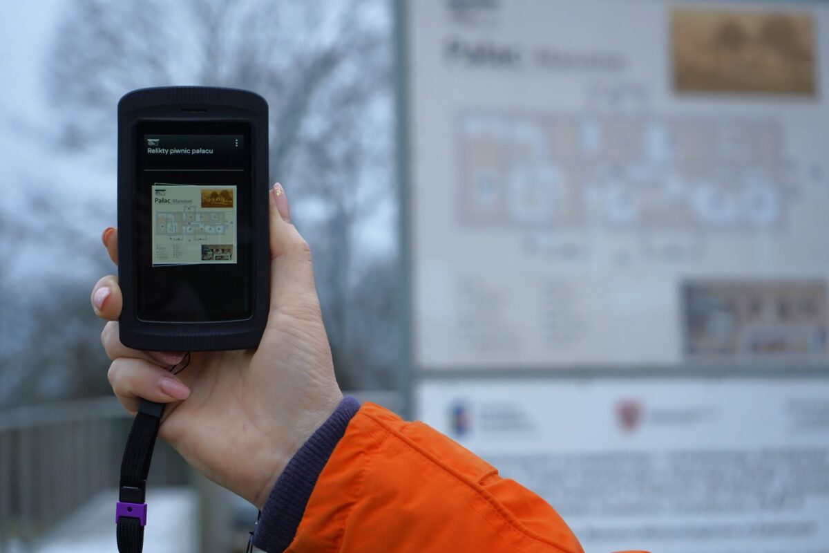 Osoba ubrana w pomarańczową kurtkę trzyma ręczne urządzenie wyświetlające niewyraźną mapę lub przewodnik, pasujące do nieostrego znaku informacyjnego w tle na zewnątrz.