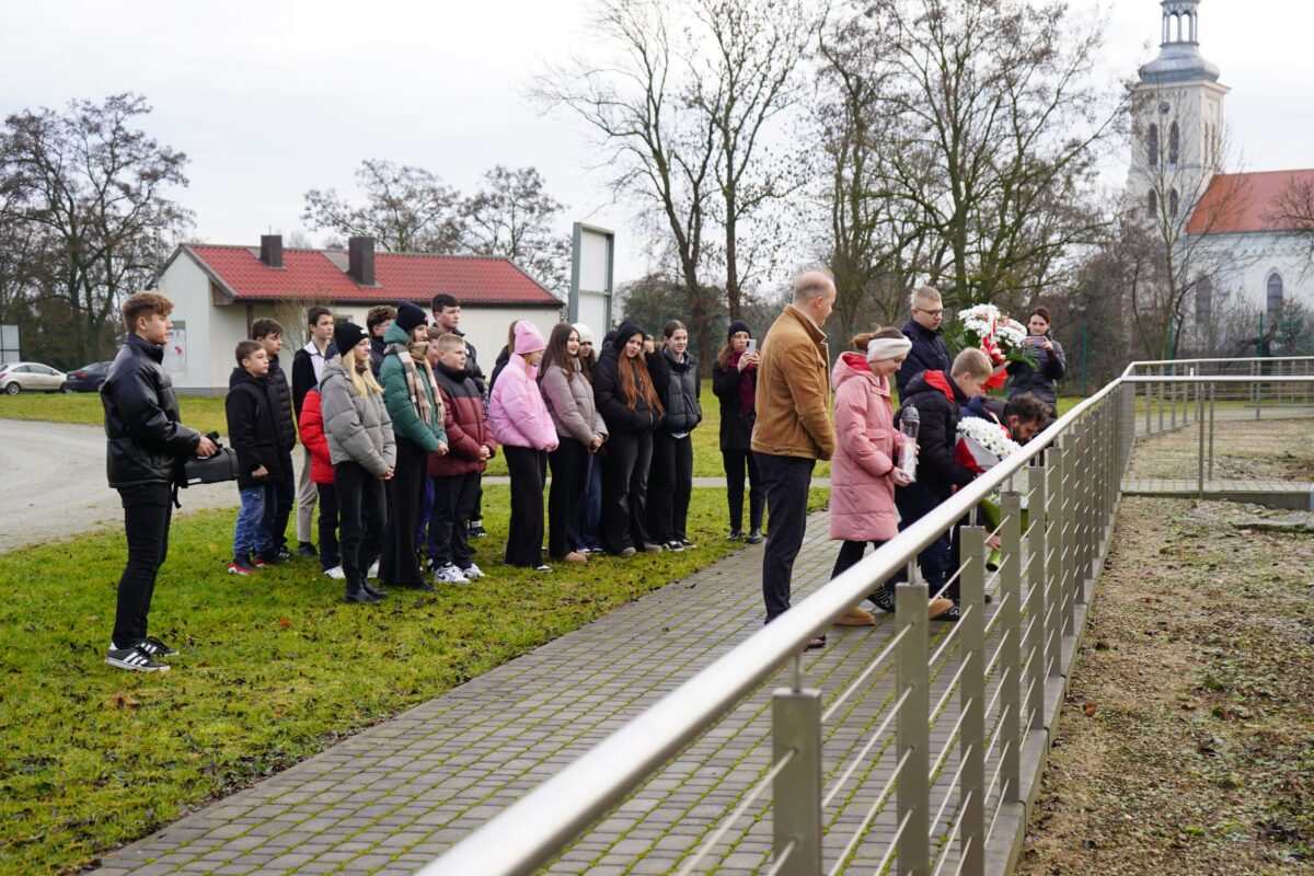 Grupa ludzi, w tym dzieci i dorośli, stoi na zewnątrz na utwardzonej ścieżce obok metalowego ogrodzenia, zwrócona w stronę trawiastego obszaru. W tle widoczne są drzewa, domy i duży kościół.