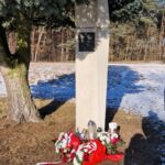 Pod drzewem stoi kamienny słup pamięci z pustą tablicą. U jego podstawy znajdują się czerwone i białe kompozycje kwiatowe, świece i wstążka. Ziemia wokół jest trawiasta z plamami śniegu, a w tle widać drzewa.