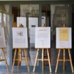 Kilka oprawionych dzieł sztuki i dokumentów wyświetlanych na drewnianych sztalugach jest ustawionych w rzędzie w pomieszczeniu, z dużymi oknami i elementami architektonicznymi widocznymi w tle.