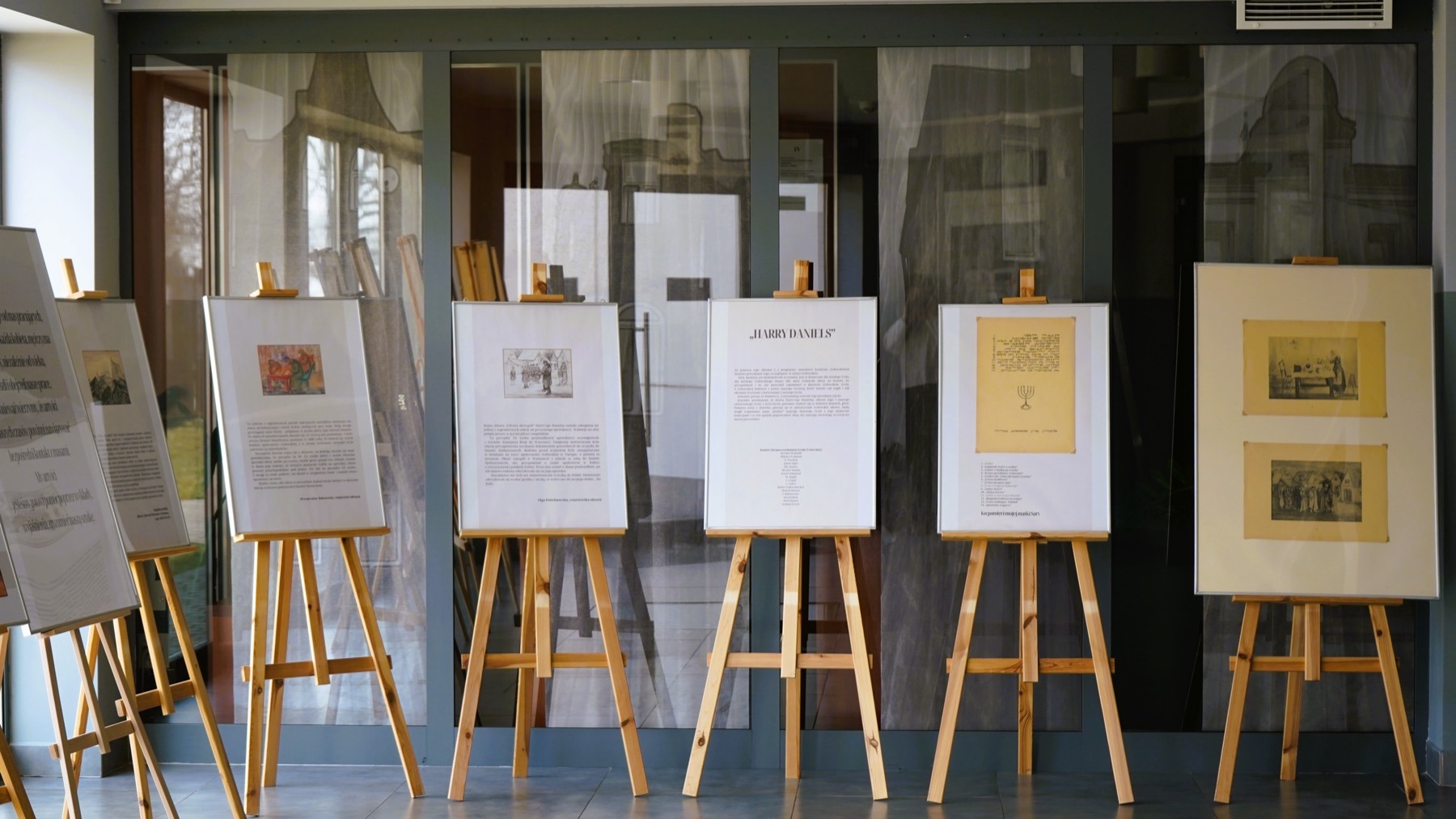 Kilka oprawionych dzieł sztuki i dokumentów wyświetlanych na drewnianych sztalugach jest ustawionych w rzędzie w pomieszczeniu, z dużymi oknami i elementami architektonicznymi widocznymi w tle.