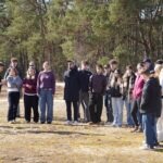 Grupa ludzi stoi na zewnątrz w słoneczny dzień w pobliżu lasu, słuchając osoby przemawiającej z przodu. Większość z nich ubrana jest w luźne, ciepłe ubrania, a ziemia pokryta jest suchą trawą.