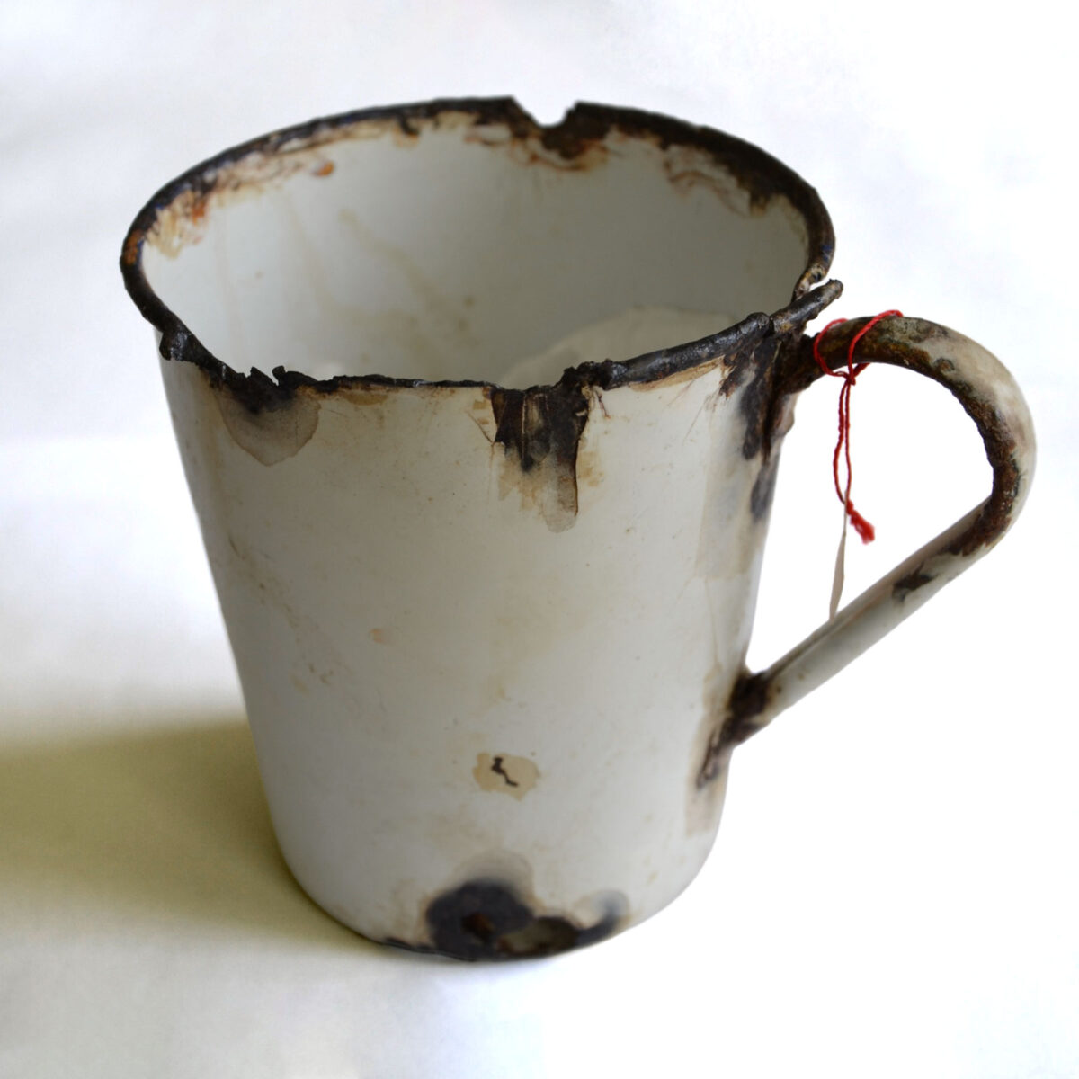 Wyszczerbiony, zardzewiały i poplamiony emaliowany metalowy kubek z uszkodzonym brzegiem i uchwytem, ustawiony na prostym białym tle. Do uchwytu przymocowany jest mały czerwony sznurek.