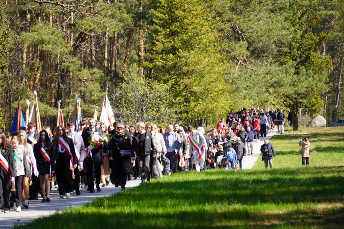 Duża grupa ludzi, niektórzy niosący flagi, idzie razem ścieżką przez zielony park otoczony drzewami w słoneczny dzień.