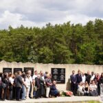 Duża grupa ludzi stoi razem przed ścianą pamięci z tablicami, otoczona drzewami pod częściowo zachmurzonym niebem. Niektóre osoby umieszczają lub układają kwiaty u podstawy ściany.