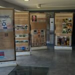 Pięć banerów informacyjnych z tekstem i obrazami rozmieszczono w dobrze oświetlonym pomieszczeniu z podłogą wyłożoną kafelkami i dużymi oknami. Niektóre plakaty przedstawiają historyczne zdjęcia i dokumenty. Po lewej stronie widoczne jest krzesło.