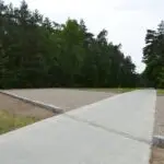 Betonowa ścieżka biegnie przez zalesiony teren, po obu stronach otoczona prostokątnymi, wyłożonymi żwirem działkami, obramowanymi kamieniami, a wszystko to w otoczeniu gęstych, zielonych drzew pod zachmurzonym niebem.