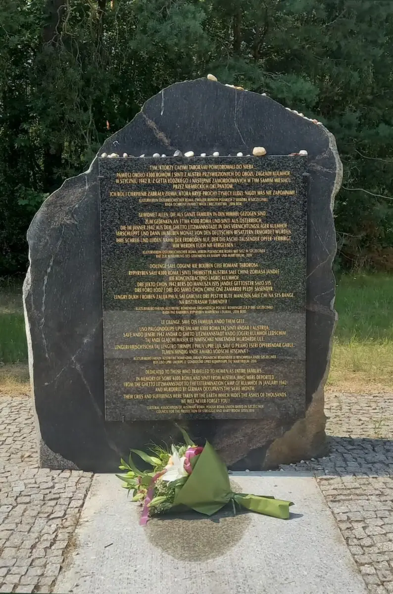 Na świeżym powietrzu, w otoczeniu drzew, stoi pomnik z czarnego kamienia z złotym napisem. Na jego szczycie leżą małe kamyczki, a u podstawy, na brukowanej ścieżce, spoczywa bukiet kwiatów.