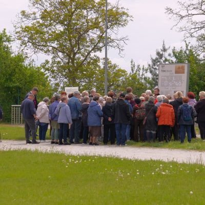 Grupa ludzi, w większości ubranych w kurtki, stoi blisko siebie na zewnątrz w pobliżu znaku informacyjnego na żwirowej ścieżce, z drzewami i zielenią w tle.