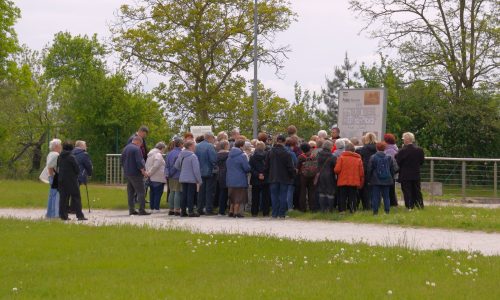 Grupa ludzi, w większości ubranych w kurtki, stoi blisko siebie na zewnątrz w pobliżu znaku informacyjnego na żwirowej ścieżce, z drzewami i zielenią w tle.
