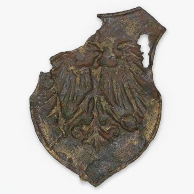 Skorodowana, uszkodzona metalowa odznaka w kształcie tarczy z wypukłym wzorem dwugłowego orła. Górna prawa część jest odłamana, odsłaniając szorstkie, postrzępione krawędzie. Powierzchnia jest nierówna i zmatowiała.