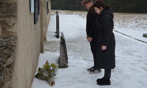 Dwie osoby stoją na pokrytej śniegiem ścieżce, pochylając głowy w geście pamięci, naprzeciwko kamiennej ściany z kwiatami ułożonymi u jej podstawy, co sugeruje moment żałoby lub hołdu w miejscu pamięci.
