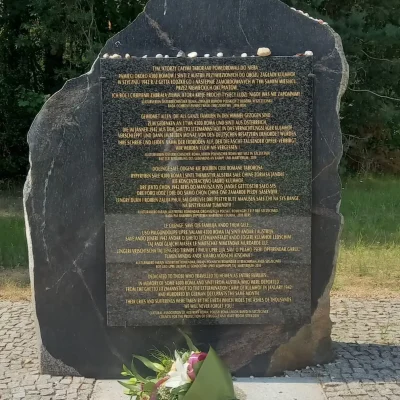 Na świeżym powietrzu, w otoczeniu drzew, stoi pomnik z czarnego kamienia z złotym napisem. Na jego szczycie leżą małe kamyczki, a u podstawy, na brukowanej ścieżce, spoczywa bukiet kwiatów.