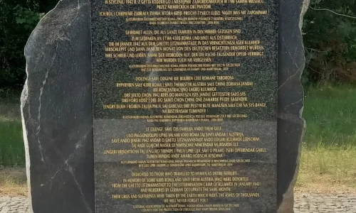 Na świeżym powietrzu, w otoczeniu drzew, stoi pomnik z czarnego kamienia z złotym napisem. Na jego szczycie leżą małe kamyczki, a u podstawy, na brukowanej ścieżce, spoczywa bukiet kwiatów.