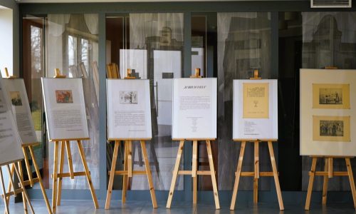 Kilka oprawionych dzieł sztuki i dokumentów wyświetlanych na drewnianych sztalugach jest ustawionych w rzędzie w pomieszczeniu, z dużymi oknami i elementami architektonicznymi widocznymi w tle.