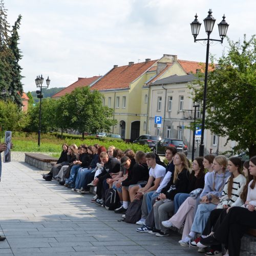 Mężczyzna stoi na zewnątrz i przemawia do dużej grupy siedzących młodych ludzi na utwardzonym terenie w pobliżu budynków i zieleni. Grupa wydaje się być uważna, a dzień jest częściowo pochmurny.