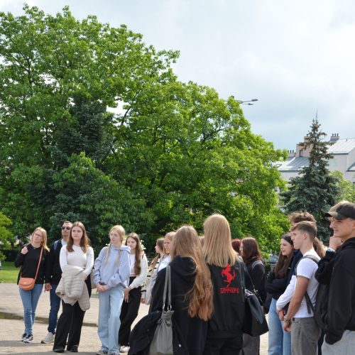 Mężczyzna stoi na zewnątrz i przemawia do grupy studentów zgromadzonych wokół niego. Grupa stoi przed zielonymi drzewami i budynkami, a nad nimi jest zachmurzone niebo. Studenci wydają się uważnie słuchać.