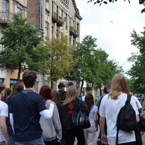Grupa ludzi idzie ulicą miejską obsadzoną drzewami, w tle widać stare budynki. Większość jest ubrana swobodnie i ma plecaki, a niebo jest zachmurzone.