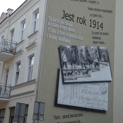 Na ścianie beżowego budynku znajduje się mural przedstawiający polski tekst, czarno-białe zdjęcie spacerujących ludzi oraz starą, ręcznie napisaną pocztówkę z 1914 r. W pobliżu widoczne są balustrady balkonów i znaki drogowe.
