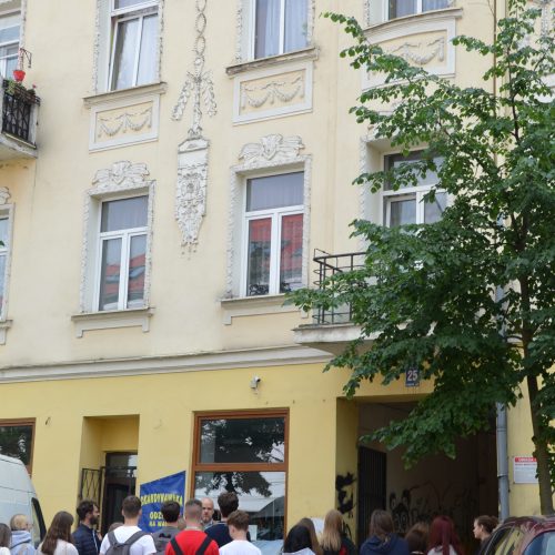 Grupa ludzi stoi na chodniku przed żółtym, trzypiętrowym budynkiem z ozdobnymi białymi wykończeniami, wysokimi oknami i drzewem częściowo zasłaniającym widok. Czerwony samochód jest zaparkowany przy krawężniku w pobliżu.