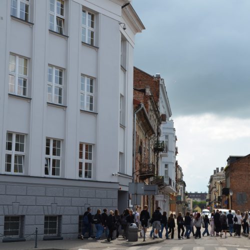 Grupa ludzi zbiera się na rogu ulicy w pobliżu dużego białego budynku z wieloma oknami pod zachmurzonym niebem. Znak drogowy na budynku głosi WŁOCŁAWKU. Inne budynki ciągną się wzdłuż ulicy w tle.