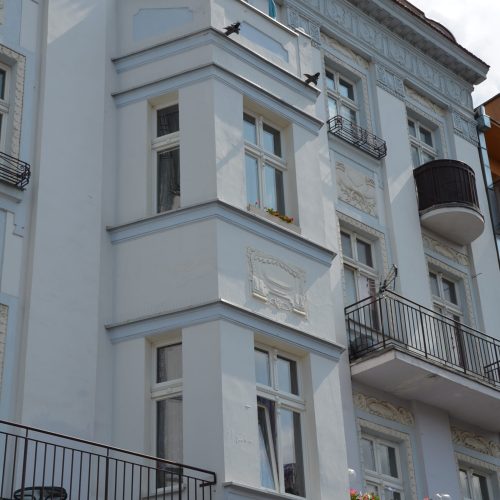 Jasnoniebieski budynek apartamentowy z ozdobnymi białymi wykończeniami, wieloma oknami i czarnymi metalowymi balkonami, niektóre z kwiatami. Niebo jest częściowo zachmurzone.