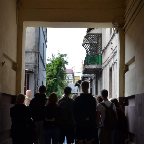 Grupa ludzi stoi w zacienionym wejściu do przejścia, patrząc w stronę jaśniejszego dziedzińca ze starymi budynkami i drzewem widocznym w tle.