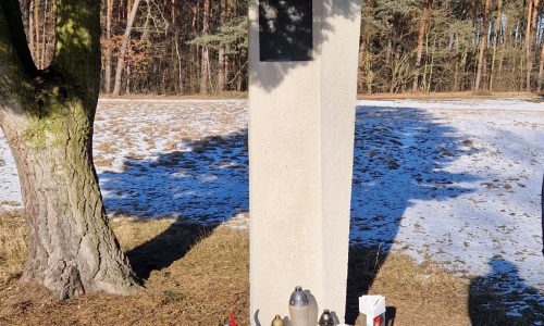 Pod drzewem stoi kamienny słup pamięci z pustą tablicą. U jego podstawy znajdują się czerwone i białe kompozycje kwiatowe, świece i wstążka. Ziemia wokół jest trawiasta z plamami śniegu, a w tle widać drzewa.