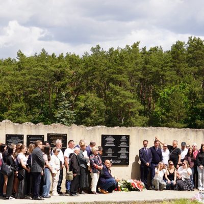 Duża grupa ludzi stoi razem przed ścianą pamięci z tablicami, otoczona drzewami pod częściowo zachmurzonym niebem. Niektóre osoby umieszczają lub układają kwiaty u podstawy ściany.