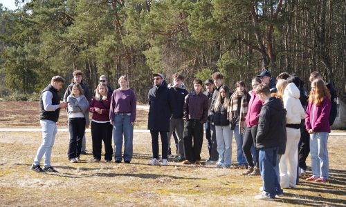 Grupa ludzi stoi na zewnątrz w słoneczny dzień w pobliżu lasu, słuchając osoby przemawiającej z przodu. Większość z nich ubrana jest w luźne, ciepłe ubrania, a ziemia pokryta jest suchą trawą.