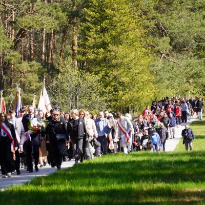 Duża grupa ludzi, niektórzy niosący flagi, idzie razem ścieżką przez zielony park otoczony drzewami w słoneczny dzień.