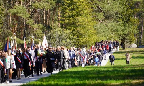 Duża grupa ludzi, niektórzy niosący flagi, idzie razem ścieżką przez zielony park otoczony drzewami w słoneczny dzień.