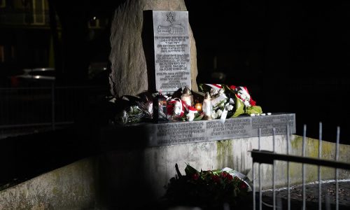 Kamienny pomnik Holokaustu w nocy, oświetlony światłem, z wieńcami kwiatowymi, zniczami i kwiatami złożonymi na pamiątkę. Tablica z inskrypcją jest częściowo widoczna na pomniku.